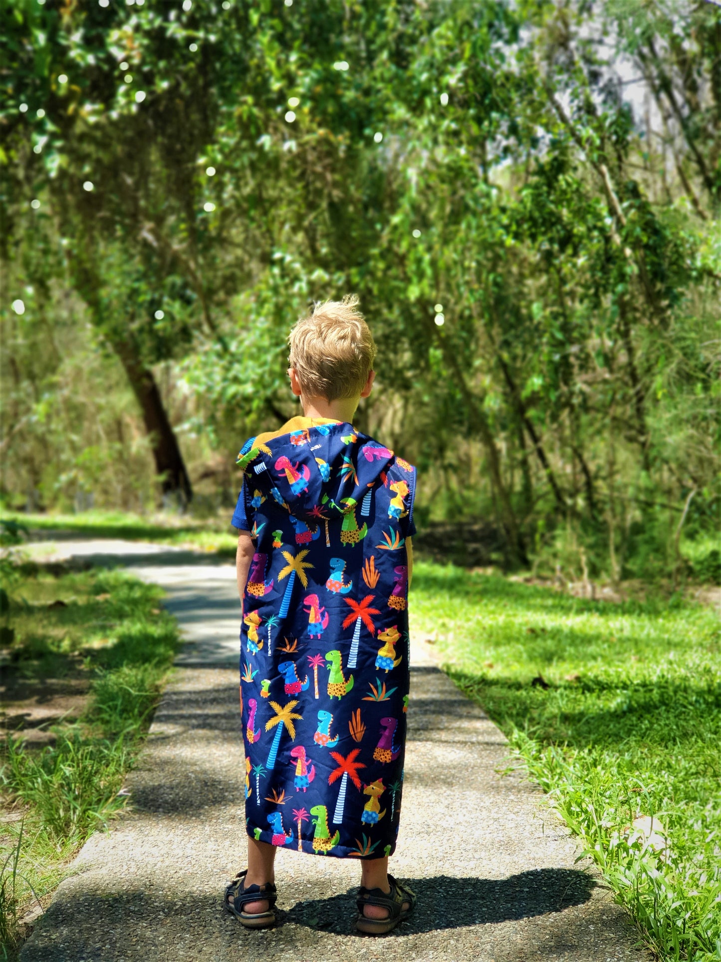 Boy wearing a size 10 swim parka. Swim parka is extra long and has dinosaur pictures on it. Bou is standing on path in currumbin beach.