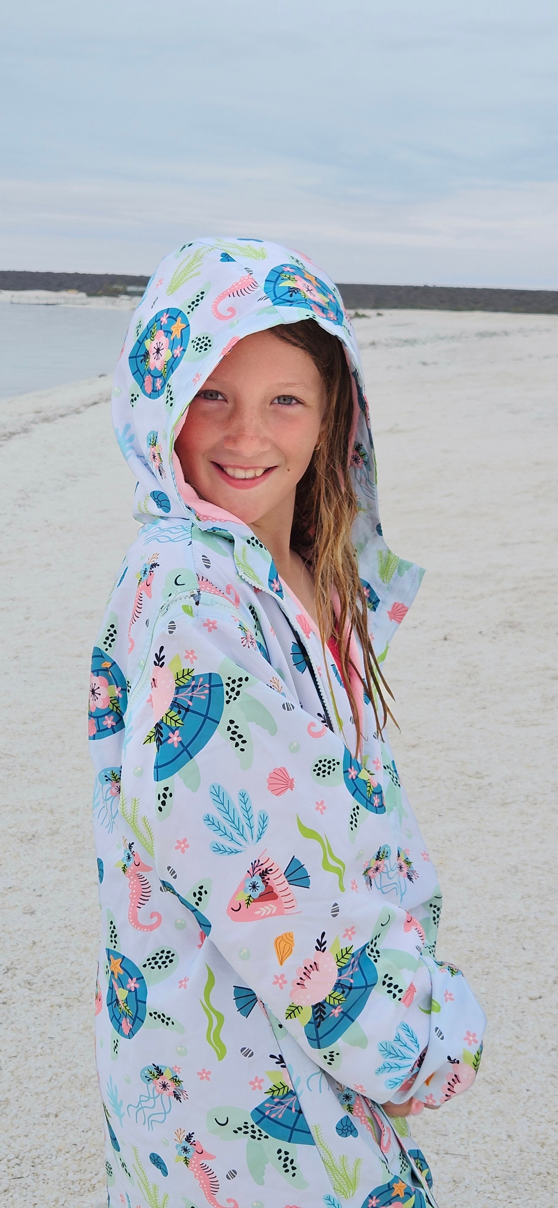Schmik Sea Haven parka with prints of turtles and sea horses worn by girl on beach. White sandy beach in background. 