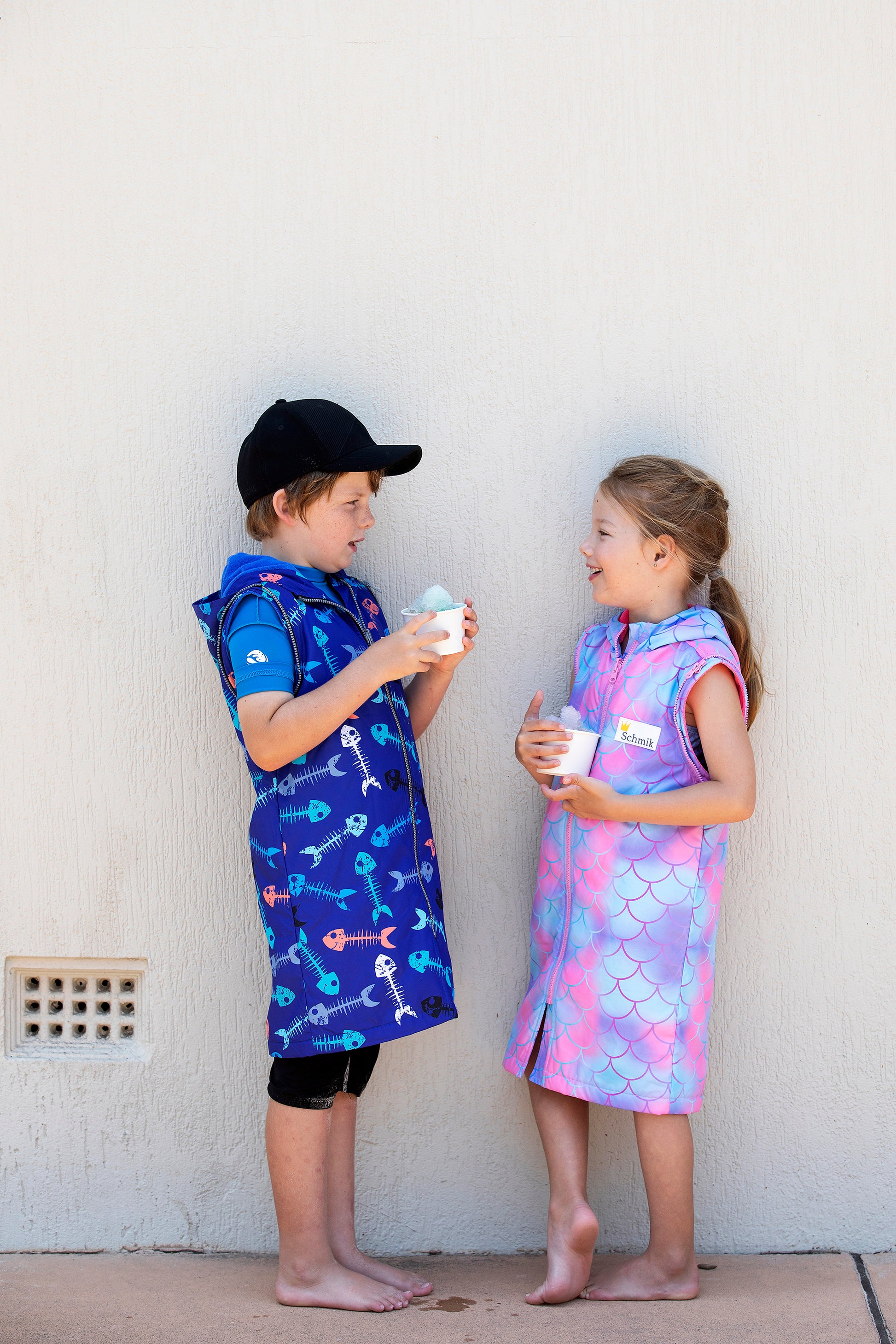 Boy standing next to girl holding a ice cup. Boy is wearing a blue fishbone print Schmik swim parka and girls is wearing a pink mermaid pattern swim parka that comes down to ther knees. 