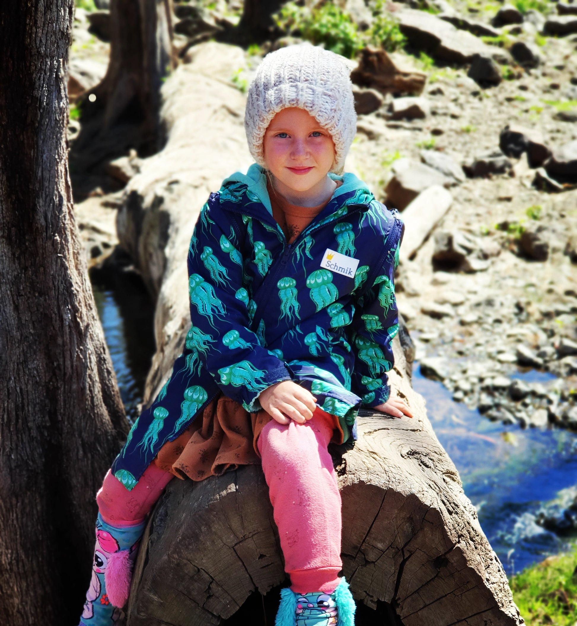 Child sitting on a log wearing a schmik swim parka. Child is camping and wears the jacket to keep warm.