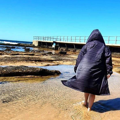 Adult wearing a black Schmik swim parka. Adult is walking on the beach and has the swim parka on with he hood on thier head. Adult is walking on the beach.
