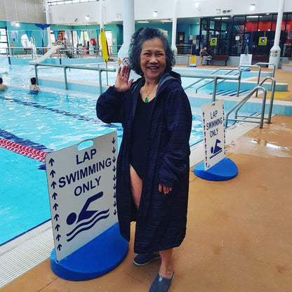 Happy asain lady wearing a black swim parka. Lady is standing in front of a lap swimming only sign. Black swim parka is long and made to be used after swimming.