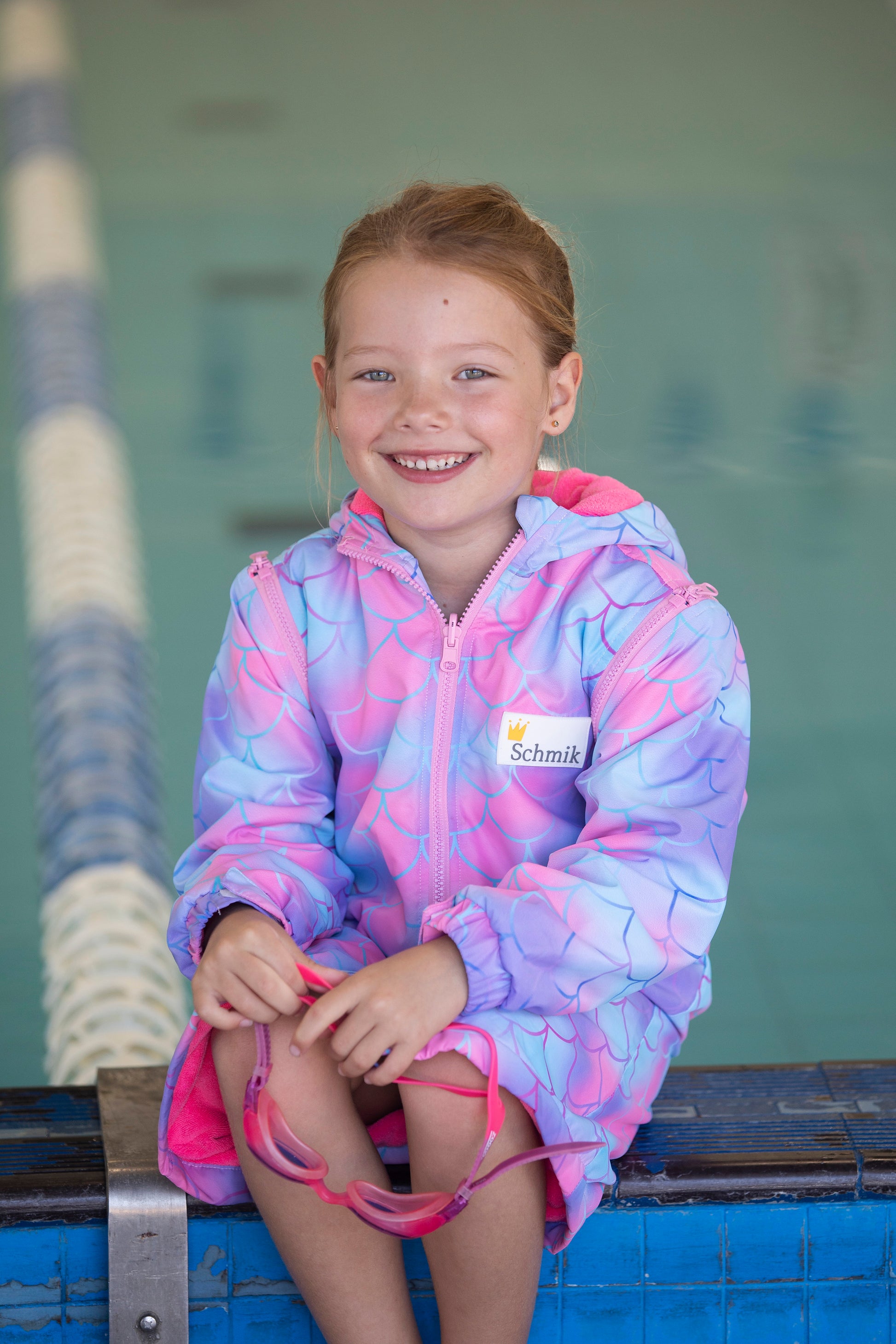 Child sitting next to pool wearing mermaid print swim parka. Mermaid print swim parka is pink and comes with removable sleeves. 