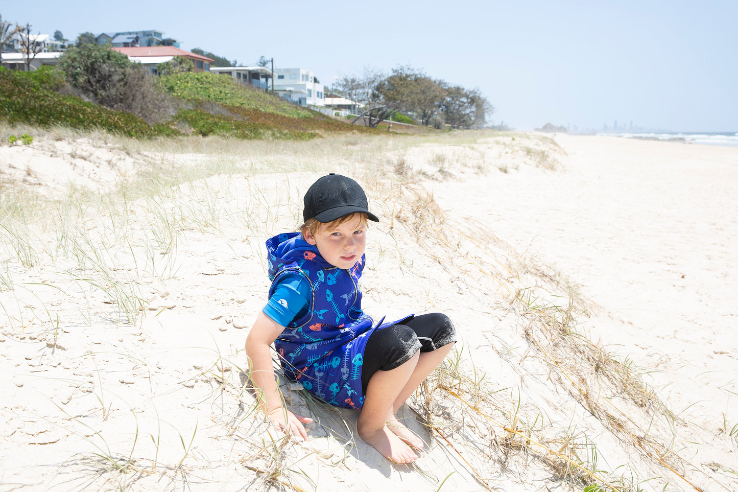 Boy sitting on the beach on the Gold Coast weaing a swim parka. Swim parka has its arms removed and is fishbone print.