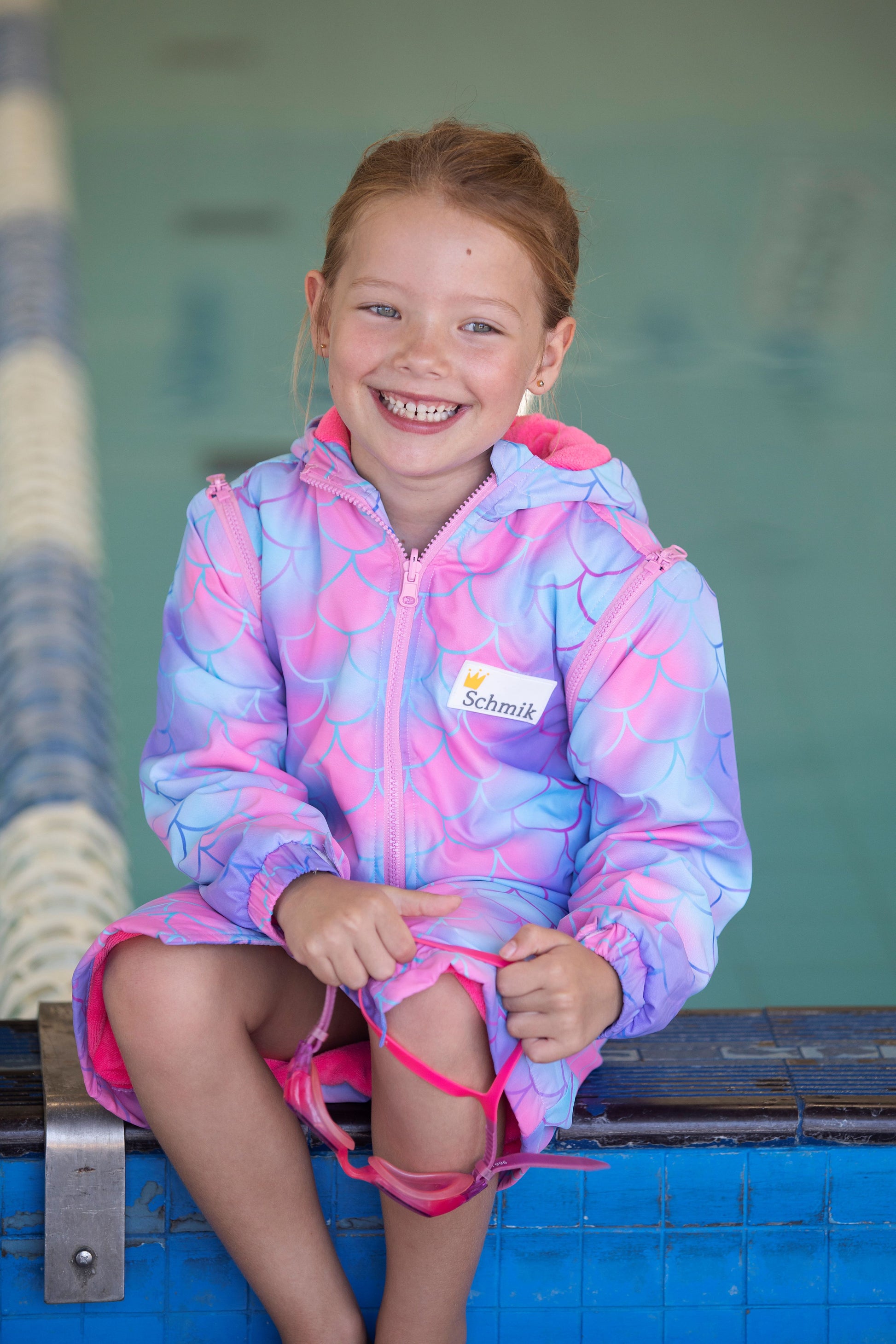 Child sitting on edge of pool. Child is wearing mermaid print swim parka and is holding goggles. Mermaid print swim parka is pink with mermaid sclales printed all over it. 
