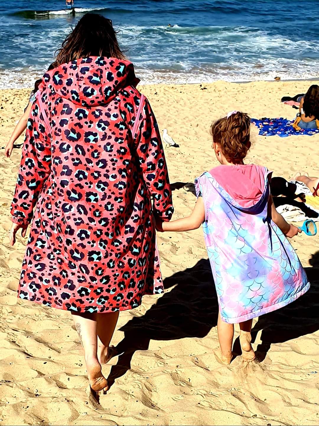Adult and child holding hands walking on the beach. Adult is wearing a animal print swim parka and child is wearing a mermaid print swim parka. 