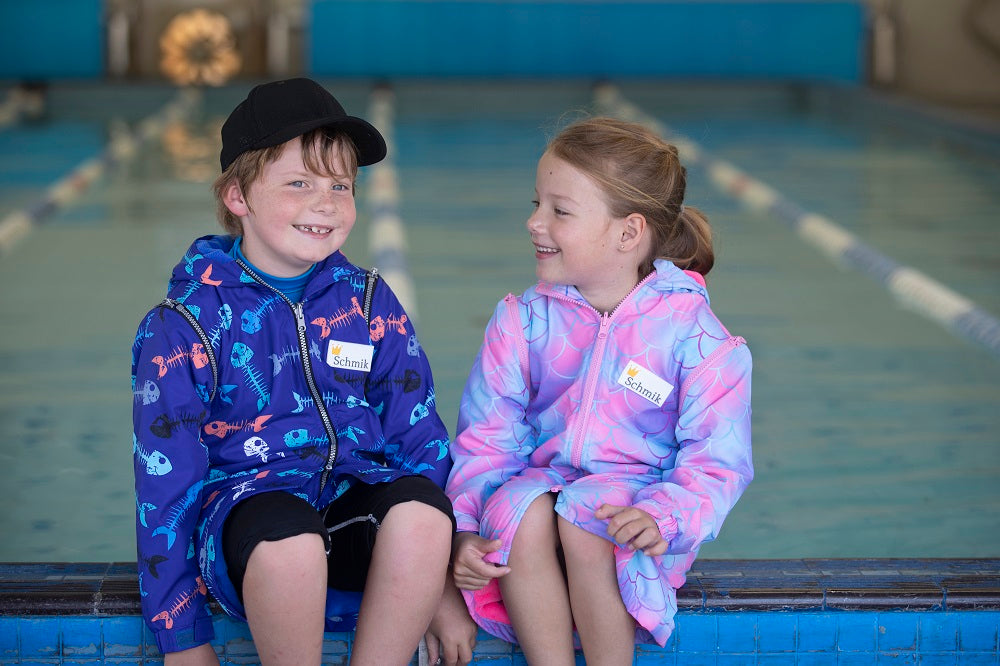 Boy and girl sitting on pool side wearing blue fishbone print swim parka and pink mermiad print swim parka. Schmik swim jackets. 
