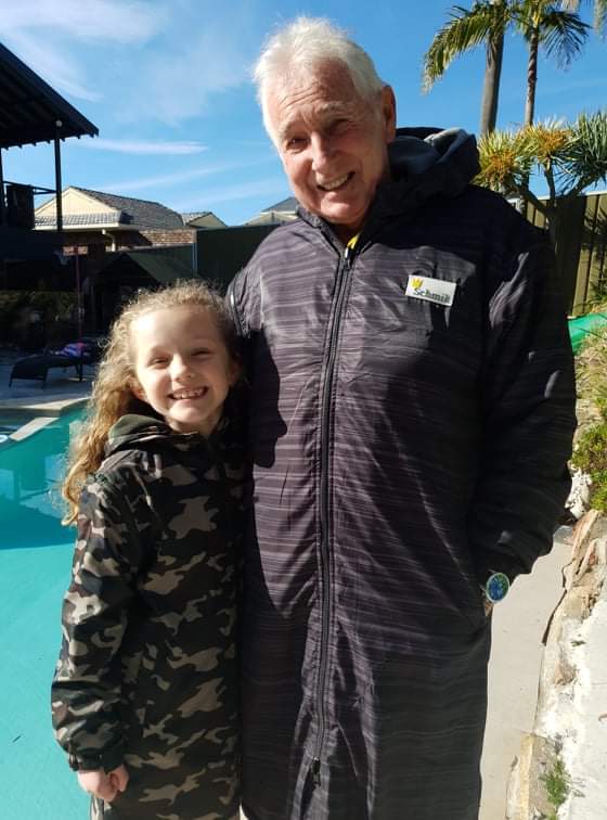 Adult and child wearing a black adult swim parka and child is wearing a kids swim parka. Both are standing standing next to each other in front of a swimming pool. Adult wears a black swim parka and child wears a army print swim parka.